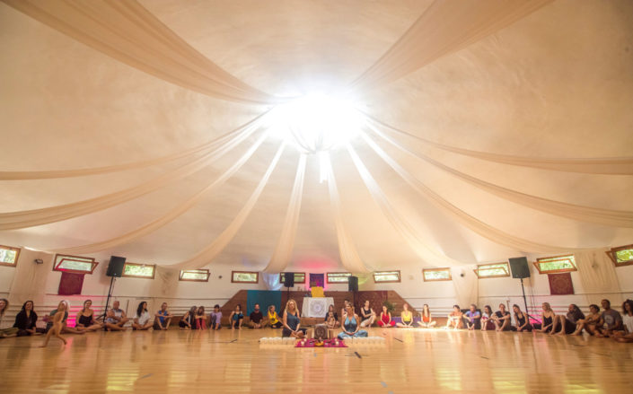 Una sala luminosa con personas sentadas en círculo. En el medio una mujer preparando una olla con cacao, a punto de servirlo para empezar una ceremonia de cacao.